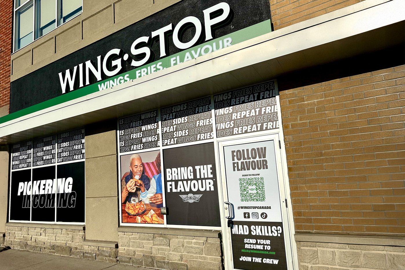 A storefront with window signage for business, having a window and two doors with prints on it. A ladder and a big can kept in front of the window of Wingstop Toronto.