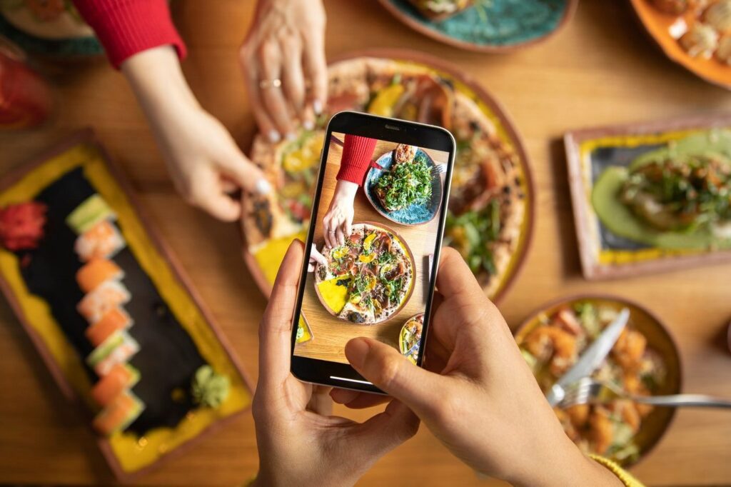 Alt text - A person holding the mobile phone in the foreground with the camera towards the table top view of various food dishes. Hands are seen in the image decorating the food dishes.